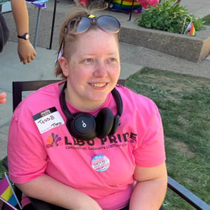 A photo of Tessa sitting in a chair at the Lebo Pride Celebration 2022 wearing a pink Lebo Pride T-shirt and their noise-cancelling headphones around their neck