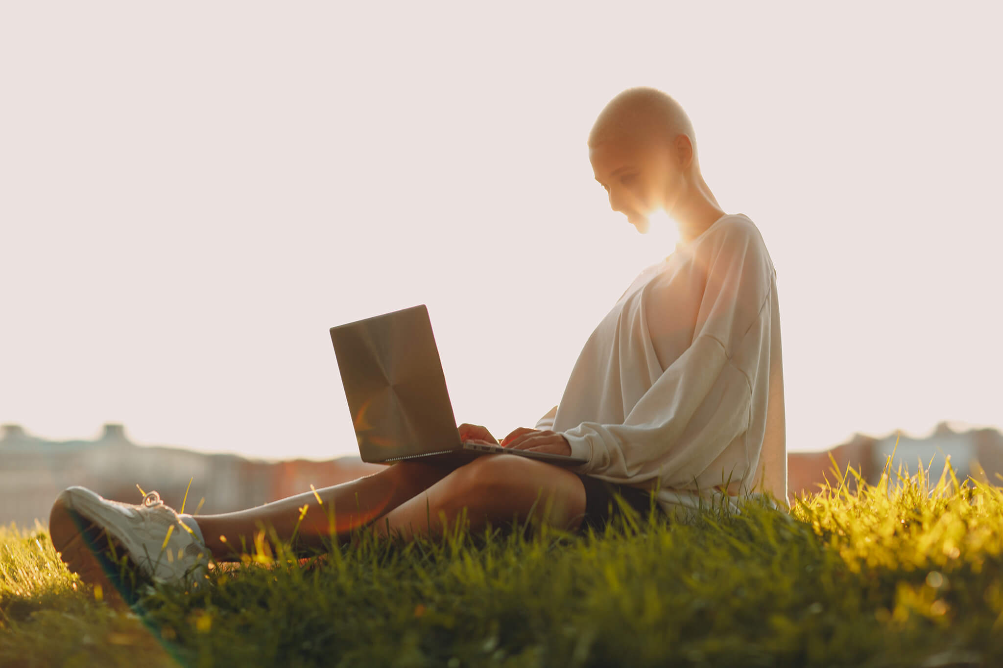 Person in white robe sitting on green grass field using silver macbook. The sun is behind their head with rays visible under their chin.