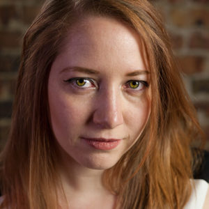 A headshot of Tessa Watkins, a white non-binary person with long, red hair and green eyes.