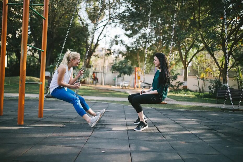 Two fem-presenting people swinging on their own swings in a playground's swingset