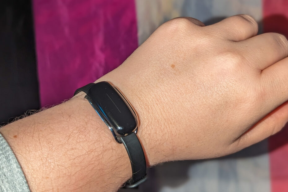 A black Bond Touch bracelet on the wrist of a white person with the genderfluid flag in the background