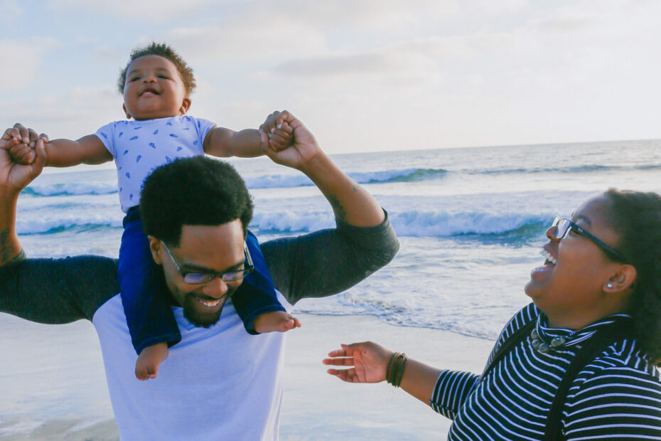 A photo of a Black family at the beach. The father is carrying their infant on his shoulders who's having a blast and the mother is laughing nearby.