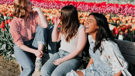 3 feminine presenting people talking and laughing while sitting on a bench in a field of flowers