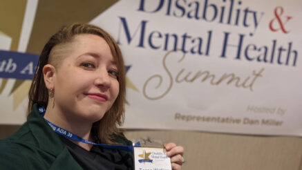 A selfie of Tessa Watkins holding up their lanyard indicating they're a speaker while standing in front of the Disability & Mental Health Summit logo