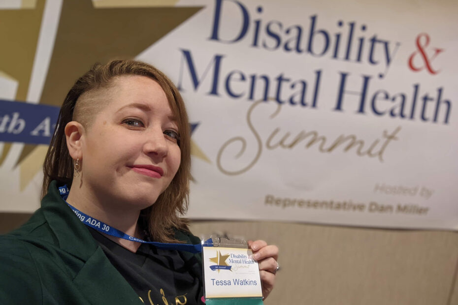 A selfie of Tessa Watkins holding up their lanyard indicating they're a speaker while standing in front of the Disability & Mental Health Summit logo