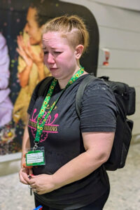 Tessa, a white autistic adult with CPTSD, is seen here standing in an airport wearing their Sunflower lanyard in distress. They are crying, stimming with their hands, and forehead is wrinkled.