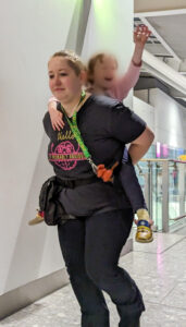 Tessa, a white autistic adult, is in an airport wearing their Sunflower lanyard, jogging about with their autistic child on their back to keep them entertained.