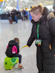 Tessa, a white autistic adult, is in an airport wearing their Sunflower lanyard while pulling their autistic child on Trunki.