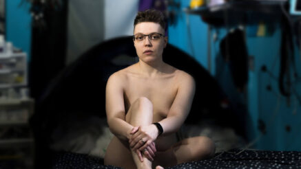 Arlo, a trans man sitting on the bed. He is naked though tastefully obscuring genitals and nipples via his pose using his arms and legs.