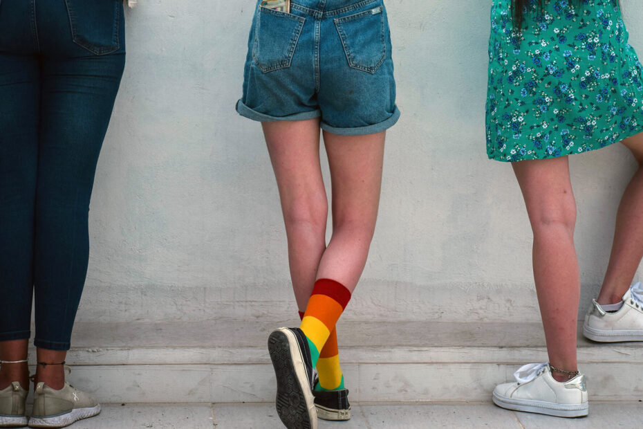 Three teens standing facing a wall. From right to left, there's a teenanger with dark skin wearing blue denim capri trousers, a white shirt, and white sneakers. The teenager in the middle is white and wearing a white shirt, blue denim shorts, black sneakers, and rainbow socks pulled up to mid-calf. The third teen has brown skin with long brown hair and is wearing a green floral dress with white sneakers.
