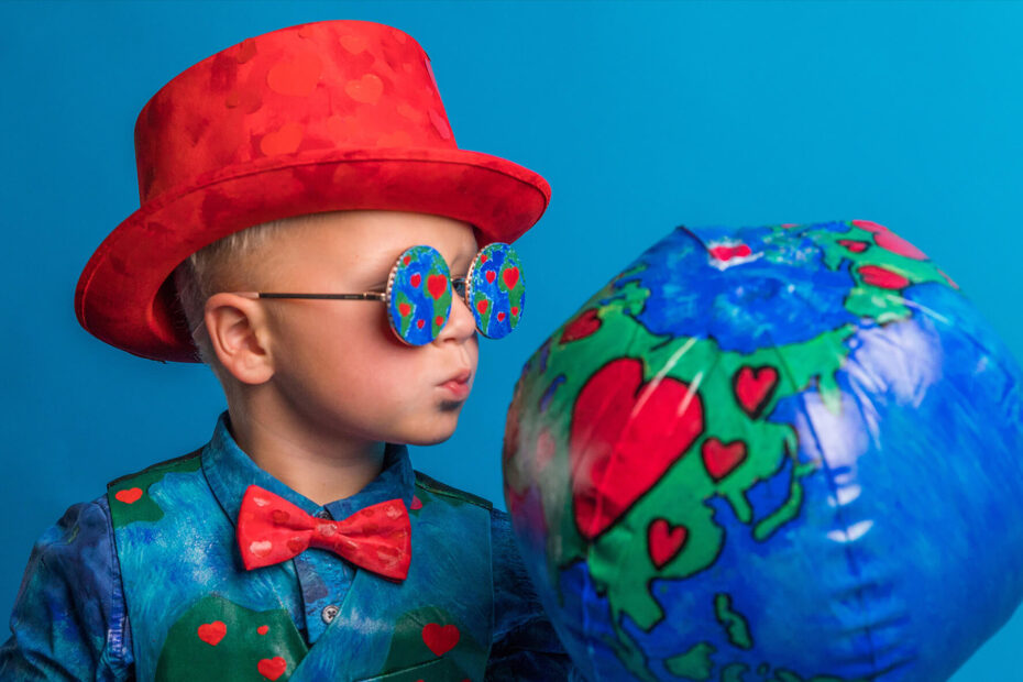 Young child in a red hat wearing a blue suit with red hearts, a red bow tie, and matching glasses, puckering up to an inflated ball that also matches the suit.