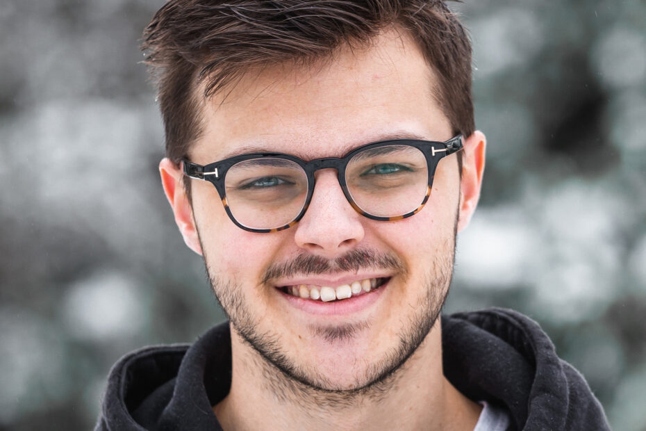 A white guy with short brown hair, black glasses, and a short facial hair wearing a black hoody smiling at the camera.