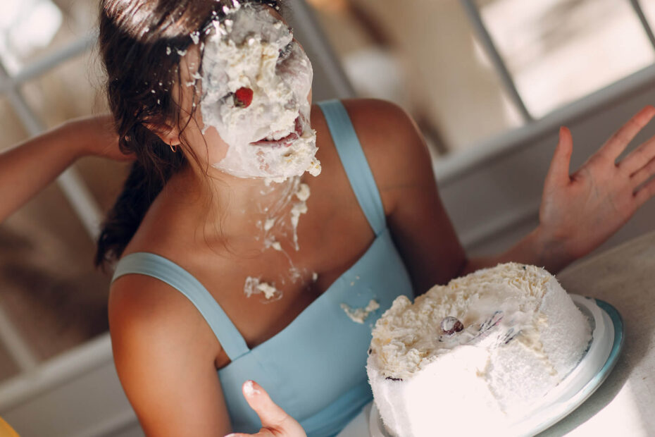 A woman in a blue dress sitting in a chair at the table with her face covered in cake after having had her face smashed into the cake sitting on the table.