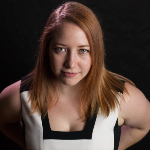 A photo of Tessa standing straight with their hands behind their back and staring at the camera. They are a white human with medium-length auburn hair wearing a predominantly white dress with black trim.