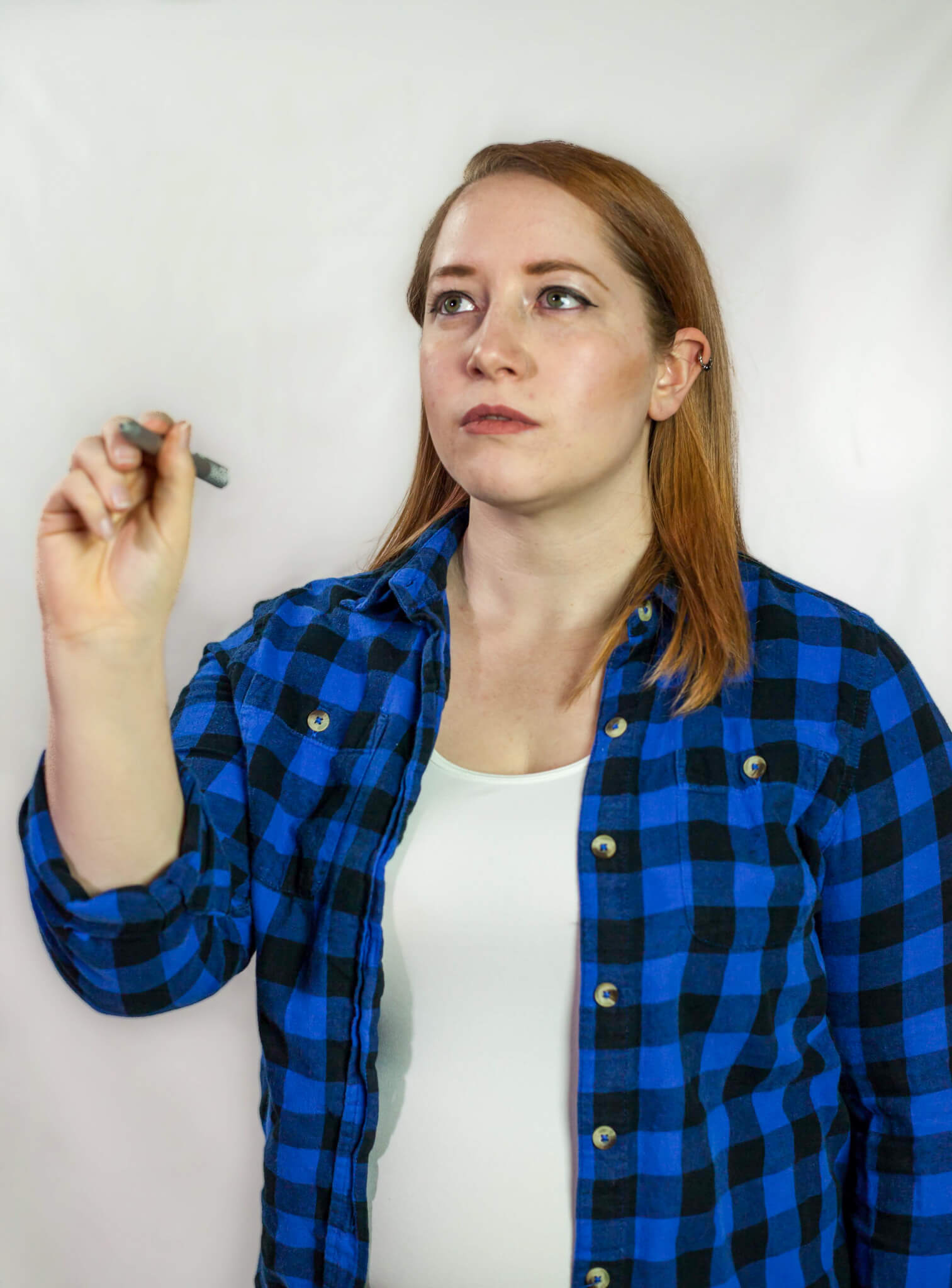 A photo of Tessa holding a stylus as if writing on a smartboard. They are a white human with medium-length auburn hair wearing a blue plaid button up shirt with a white camisole underneath.