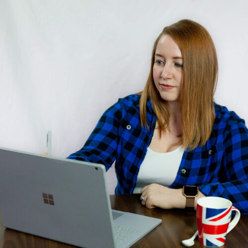 A photo of Tessa working at a laptop. They are a white human with medium-length auburn hair wearing a blue plaid button up shirt with a white camisole underneath.